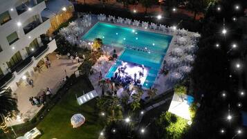 An outdoor pool area with a stage for events, surrounded by white tables and chairs, and a small fountain.