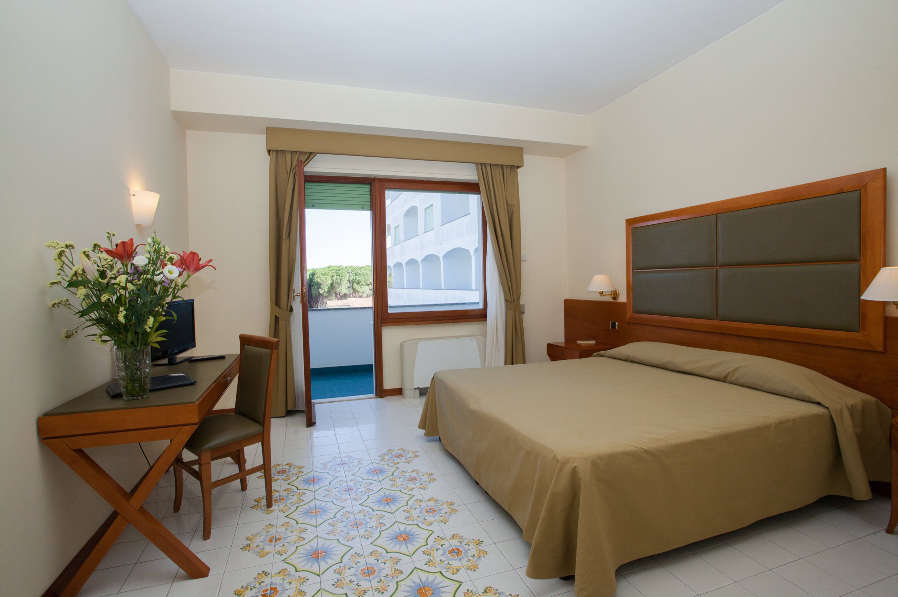 Habitación de hotel con cama, escritorio con silla, un florero, una lámpara y vista a un edificio exterior a través de una ventana.