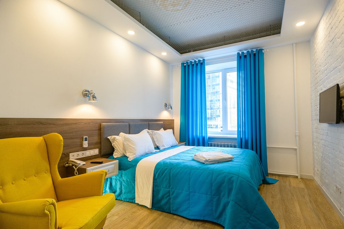 A hotel room with a bed, a yellow armchair, a nightstand, a TV, and a window with blue curtains.