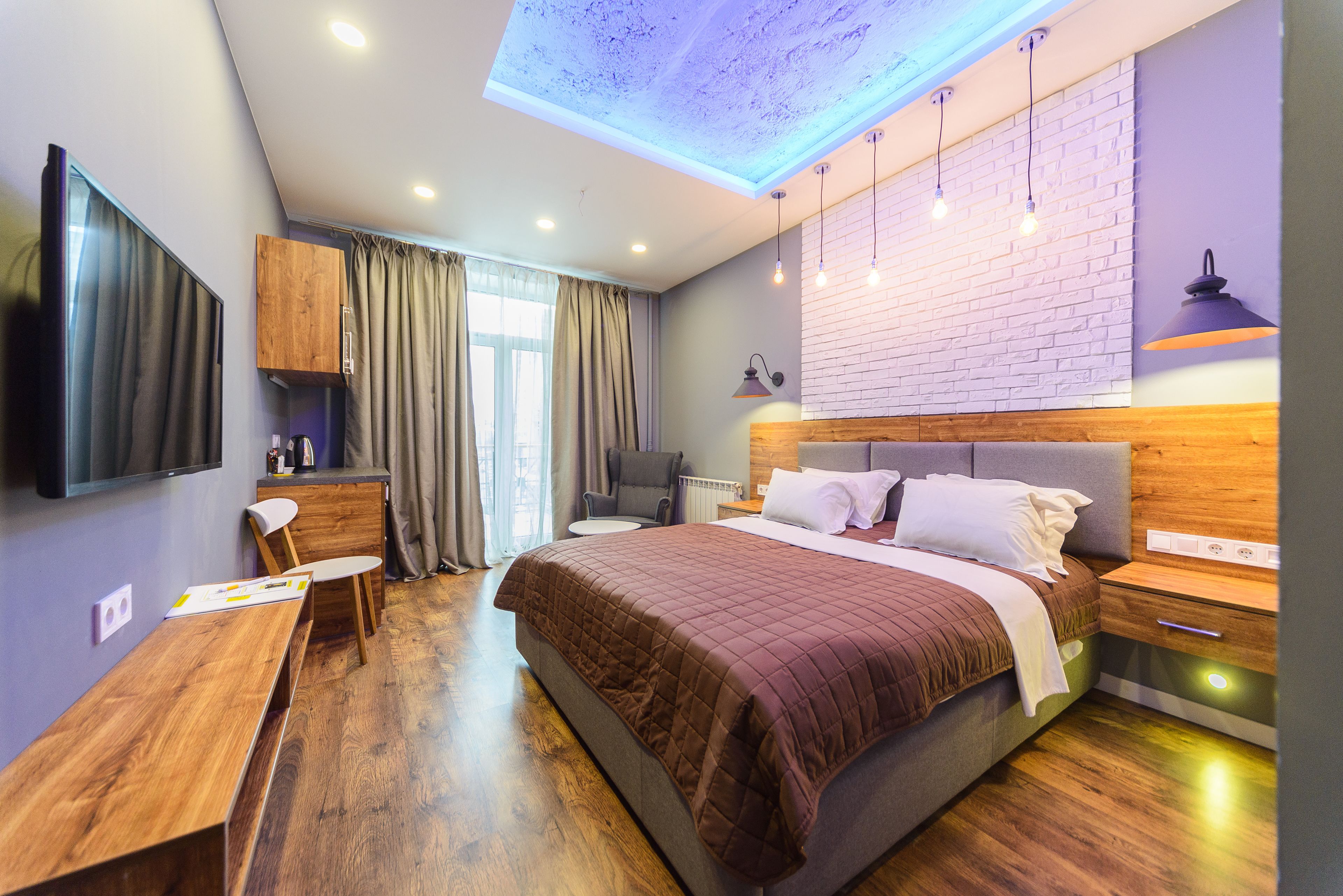 A modern bedroom with a large bed, a desk, a chair, a TV, and a ceiling light.