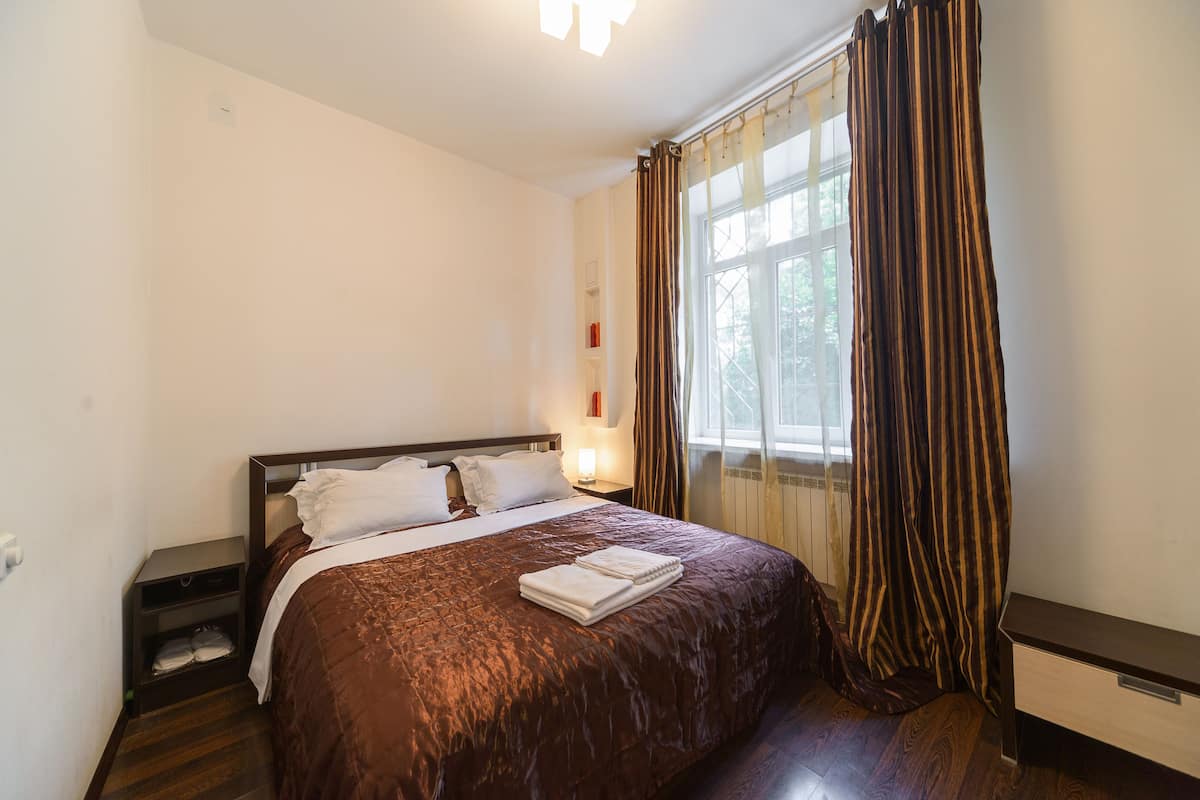 A hotel room with a large bed, a nightstand, a window with curtains, and a wooden floor.