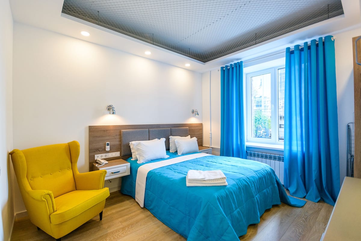 A hotel room with a bed, a yellow armchair, a nightstand, and a window with blue curtains.