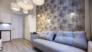 En moderne stue med en grå sofa, et lille bord og en væg med tropisk tapet. Der er tre hængende lamper og en hvid dør i baggrunden.