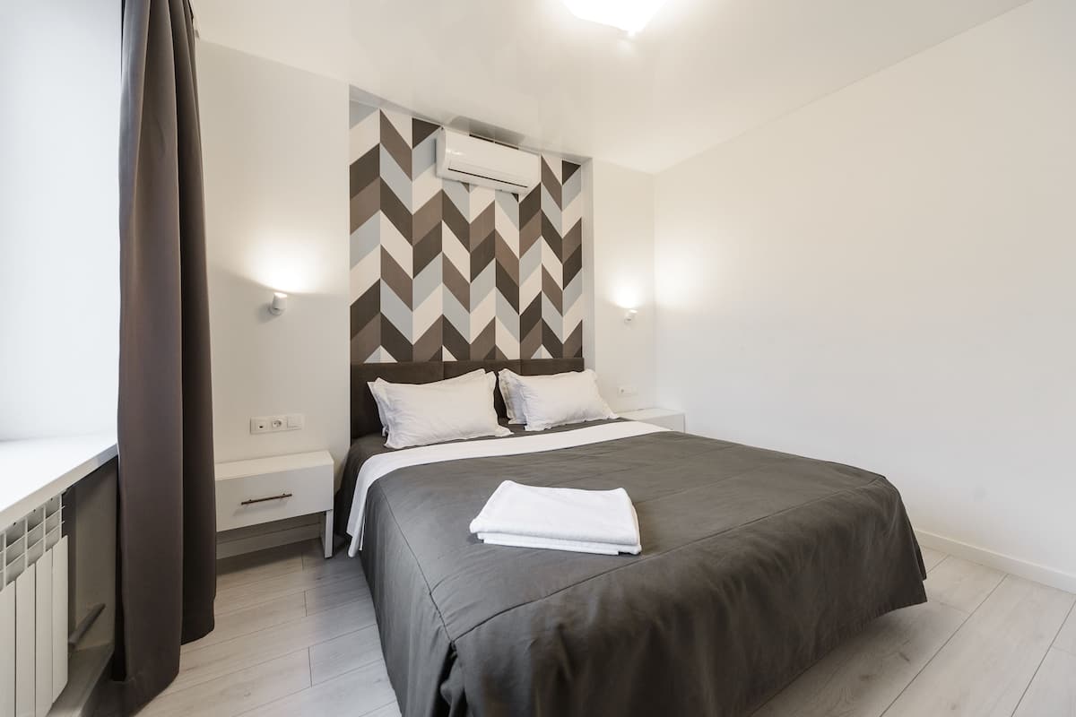A modern bedroom with a large bed, a headboard with a geometric pattern, a bedside table, and a wall-mounted air conditioner.
