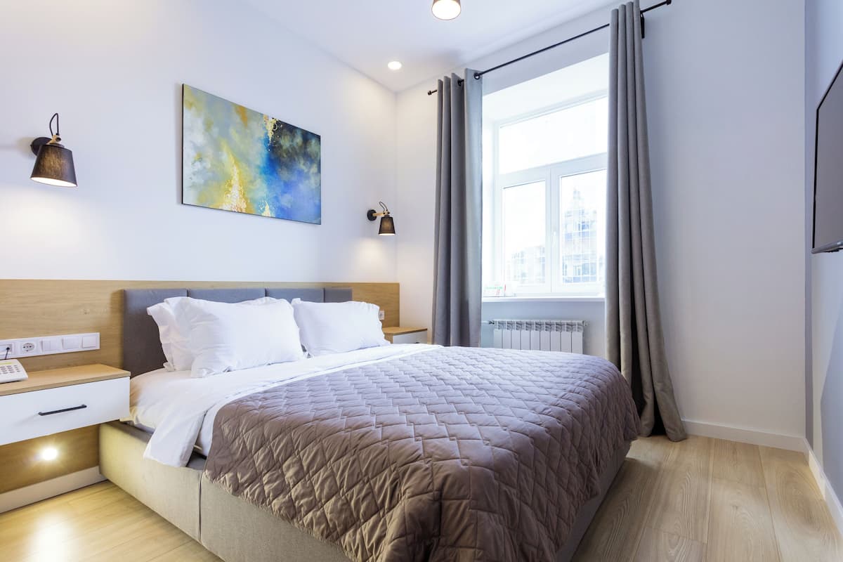 A modern hotel room with a large bed, a nightstand, a wall-mounted lamp, and a window with curtains.