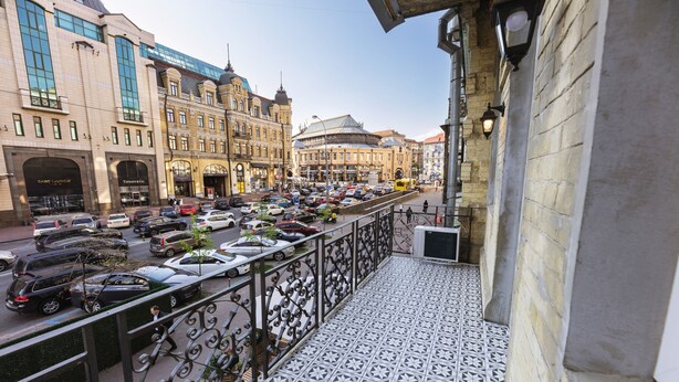 En balkon med et mønstret gulv, der kigger ud over en travl gade med parkerede biler og gamle bygninger.