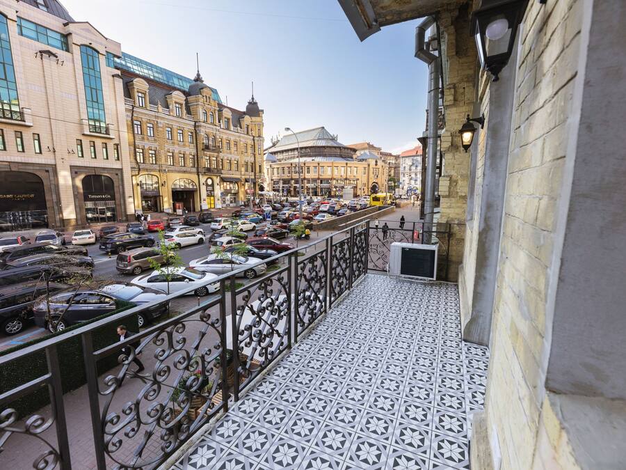Un balcón con piso estampado, con vista a una calle concurrida, autos estacionados y edificios históricos.
