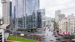 Bybillede med moderne, høje bygninger, en travl gade fyldt med biler og fodgængere, og en markant glasbygning der afspejler den omkringliggende arkitektur.