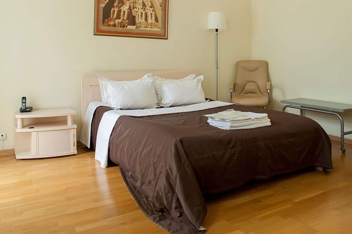 A bedroom with a bed, bedside table, lamp, chair, and a painting on the wall.
