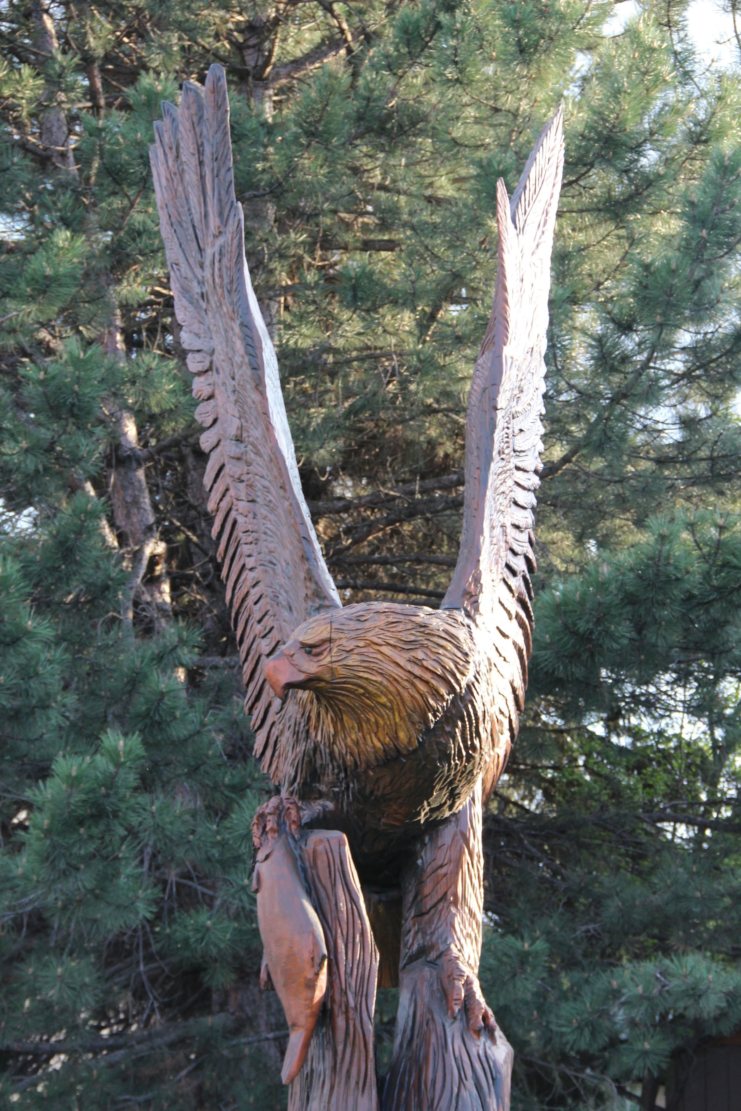 A sculpture of a bird with outstretched wings, set against a backdrop of pine trees.