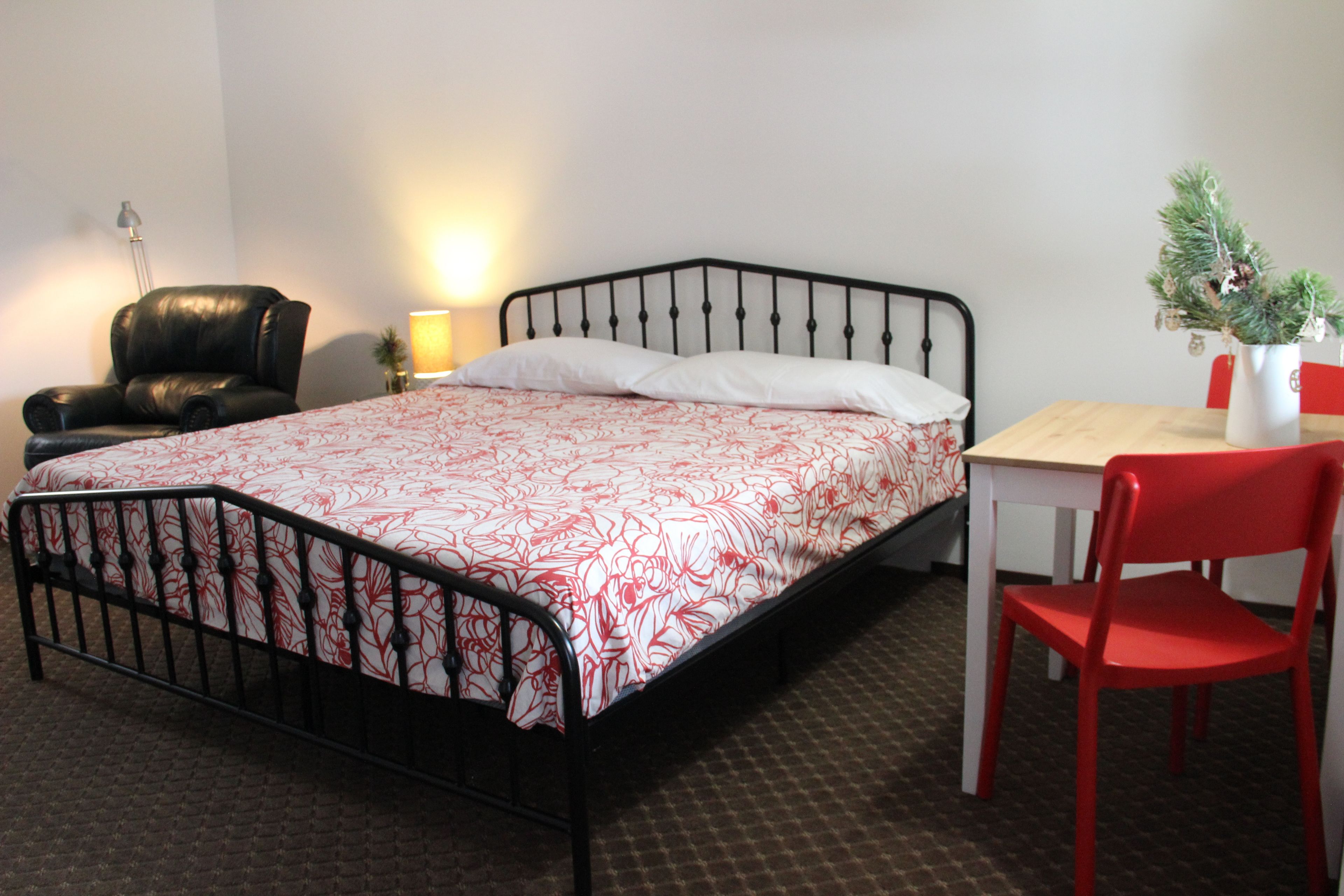 A single bed with a metal frame, a bedside table, a red chair, a lamp, and a small plant on the table.