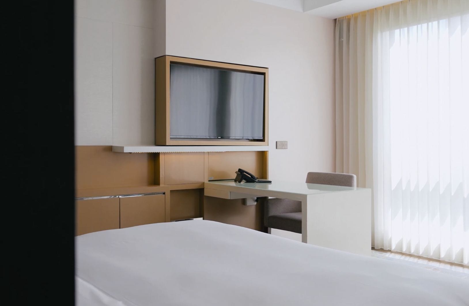 A hotel room with a bed, a desk with a telephone, a television mounted on the wall, and a window with curtains.
