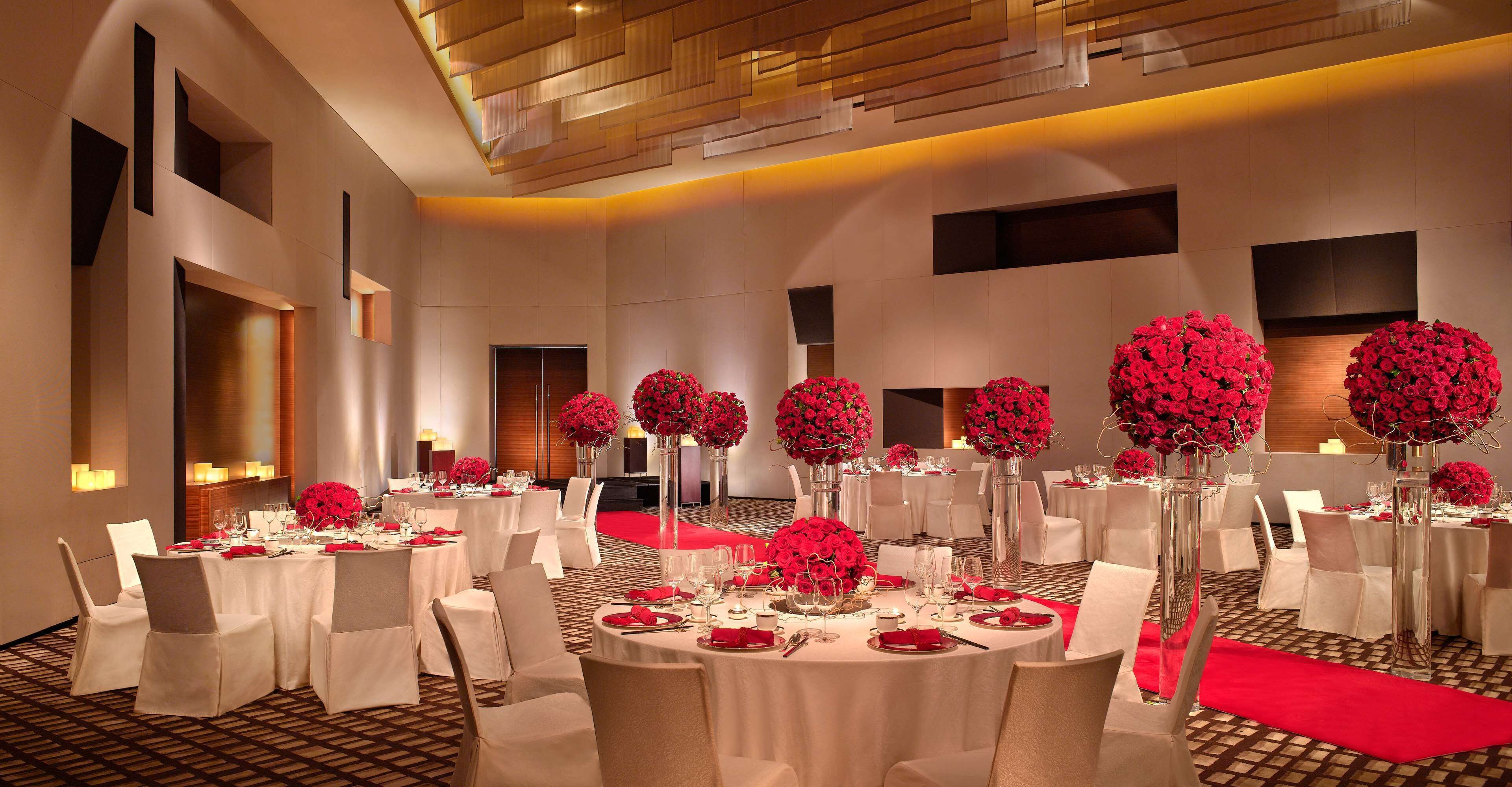 Salão de banquetes com mesas redondas, cadeiras brancas e arranjos florais vermelhos.