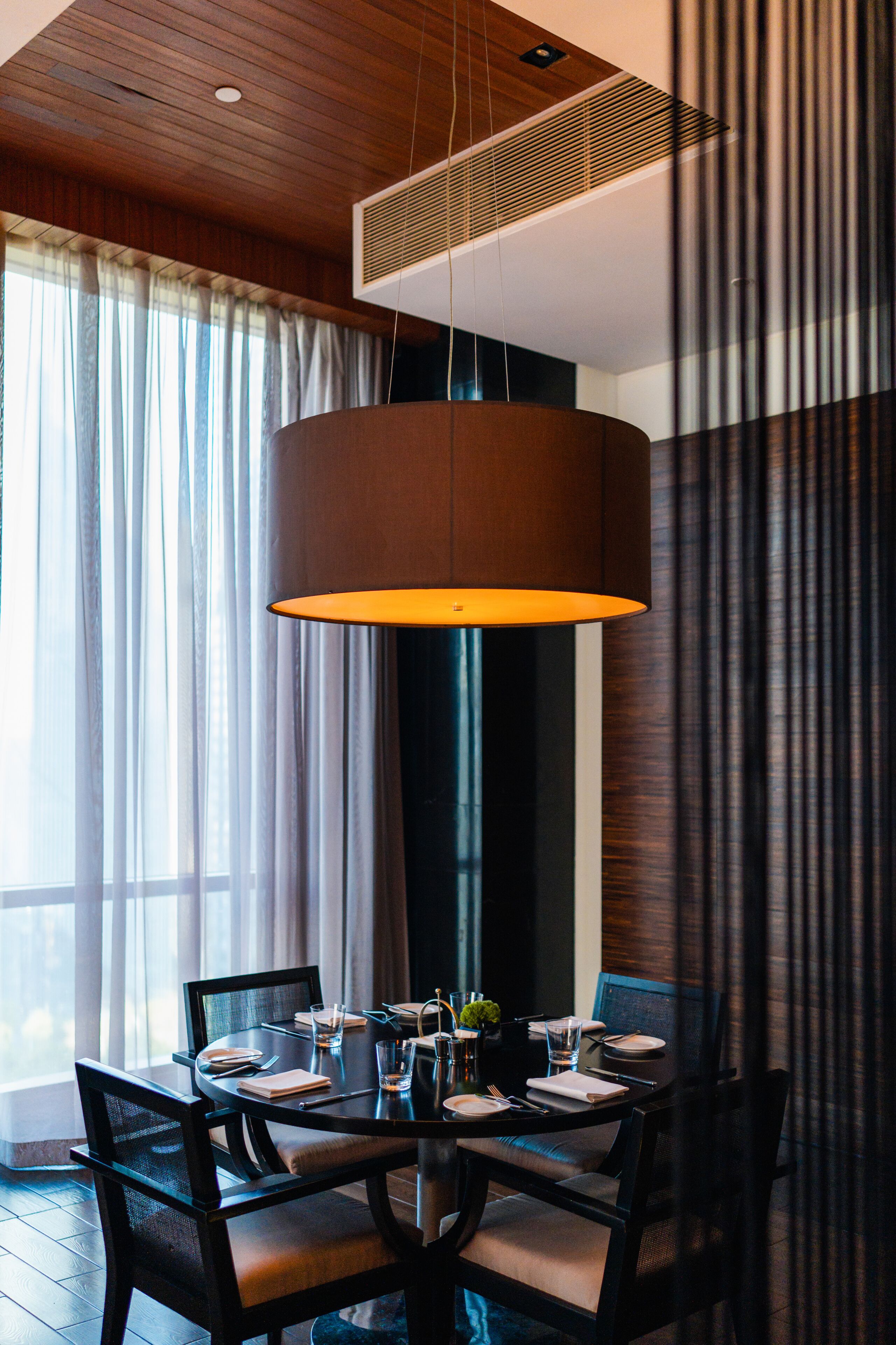 A dining area with a round table set for four, a hanging pendant light, and a view of the outdoors through large windows.