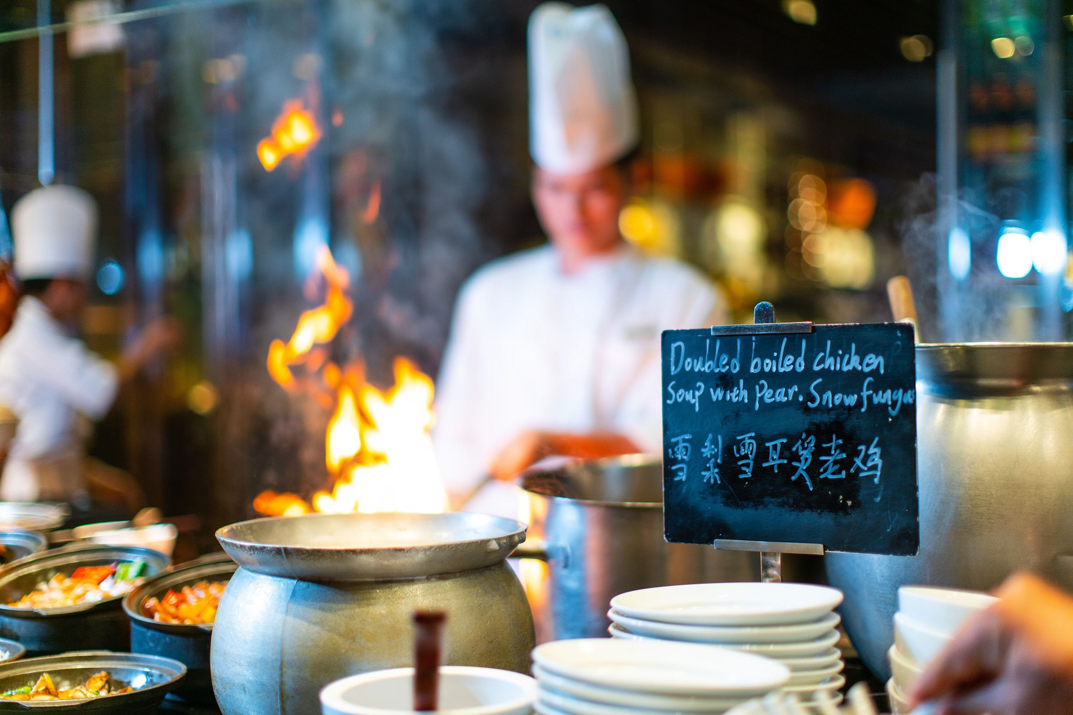 A kitchen with pots and pans on the stove, a chef in the background, and a menu board displaying dishes.