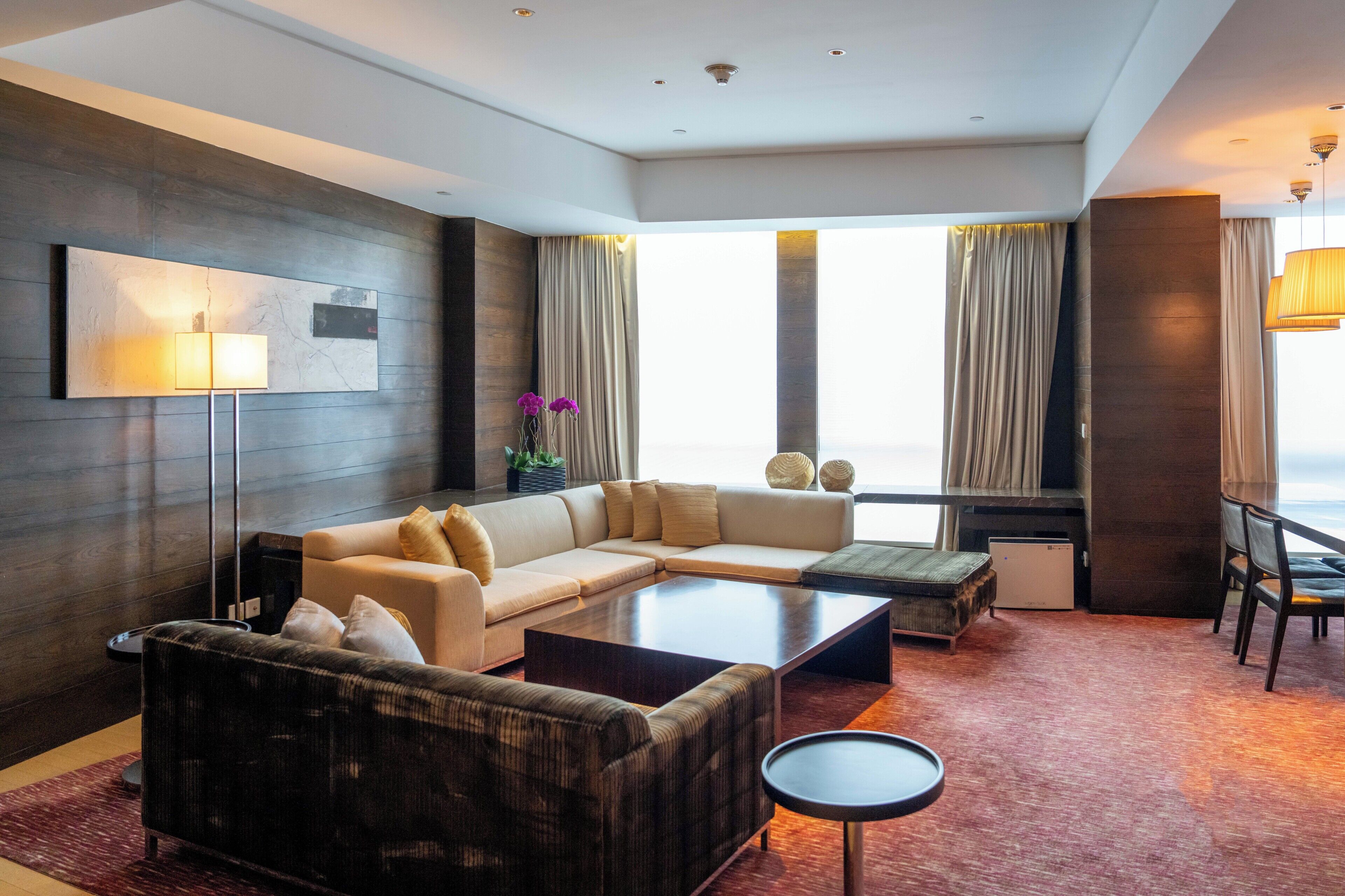 A modern hotel room with a sectional sofa, a small coffee table, a flat-screen TV, a mini-fridge, and a large window with curtains.