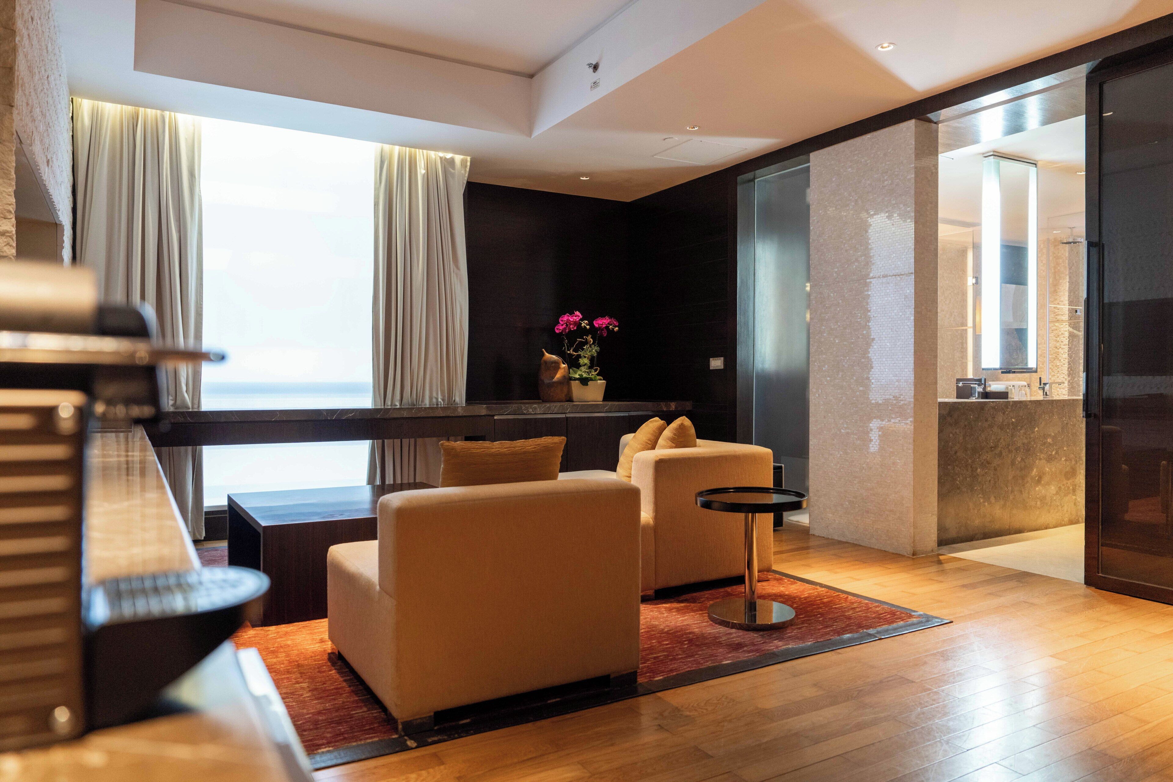 Una habitación de hotel moderna con sofá, una mesita, un jarrón decorativo con flores y vistas al mar a través de amplios ventanales.