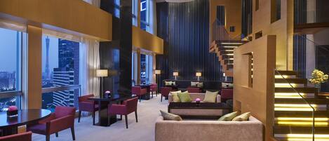 A spacious hotel lobby with a high ceiling, large windows offering city views, a central seating area with couches and chairs, and a staircase with illuminated steps.