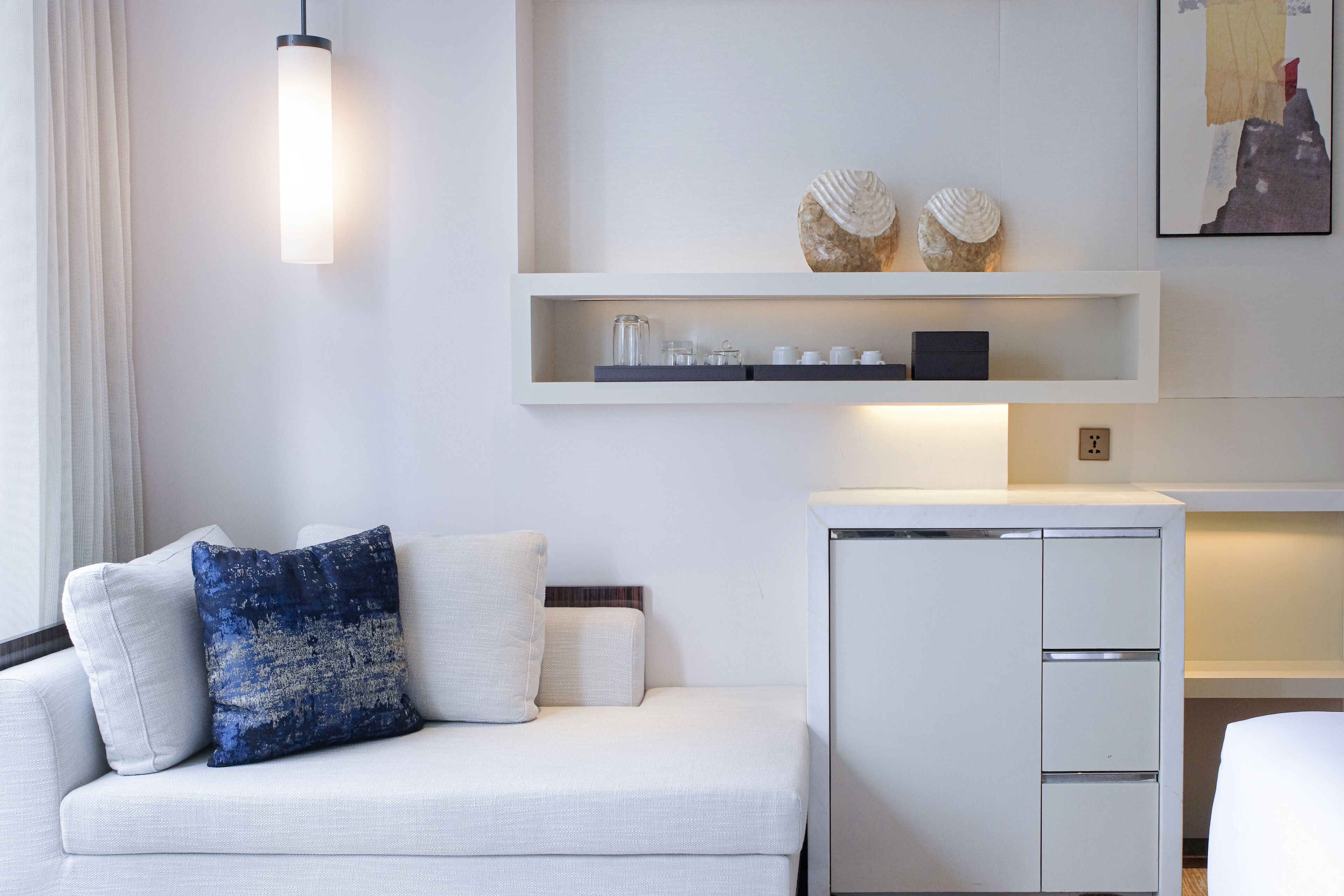 Een moderne hotelkamer met een witte bank, een klein wit kastje en een wandplank met decoratieve objecten.