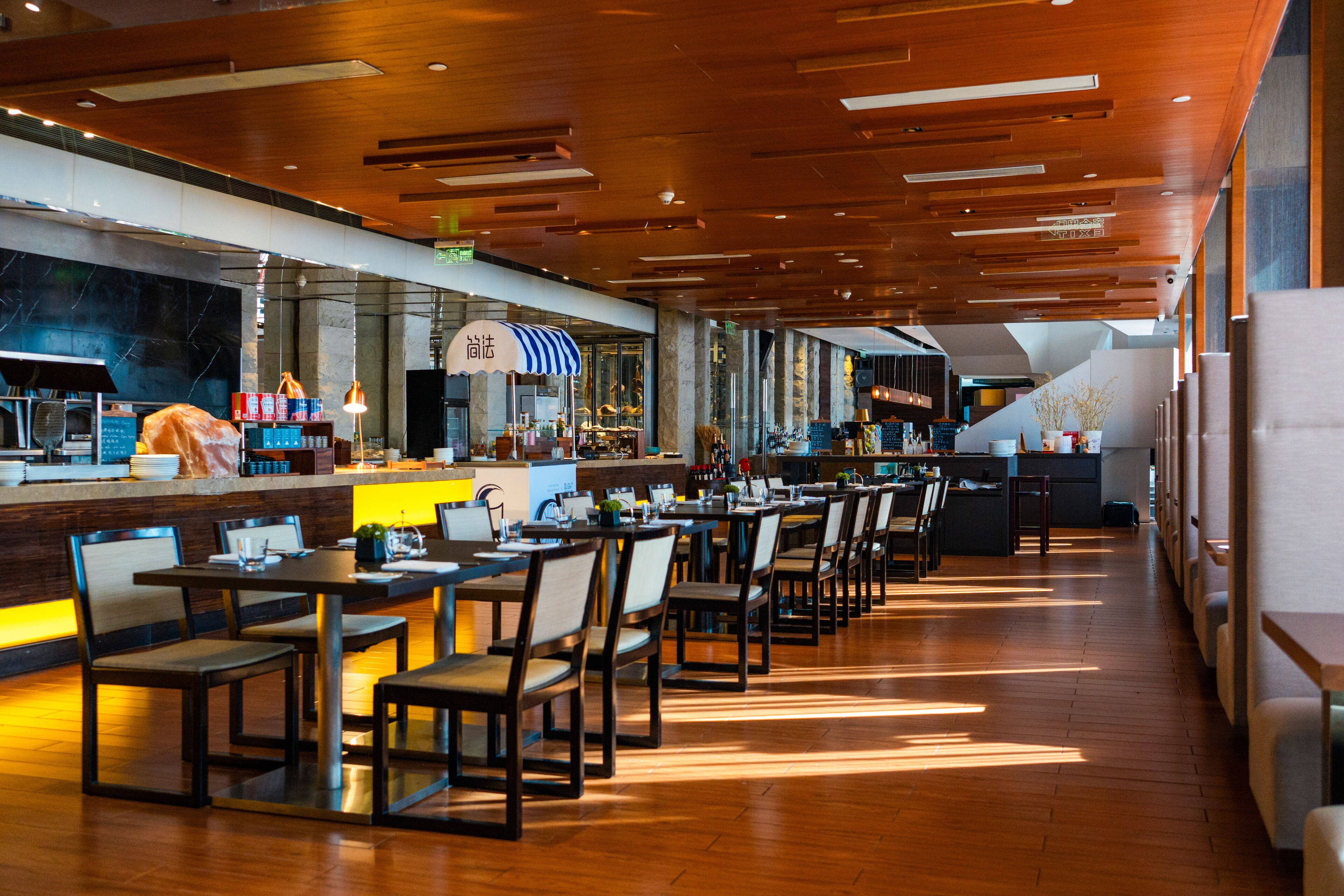 A modern restaurant with wooden flooring, a long counter with a striped umbrella, and multiple tables and chairs.