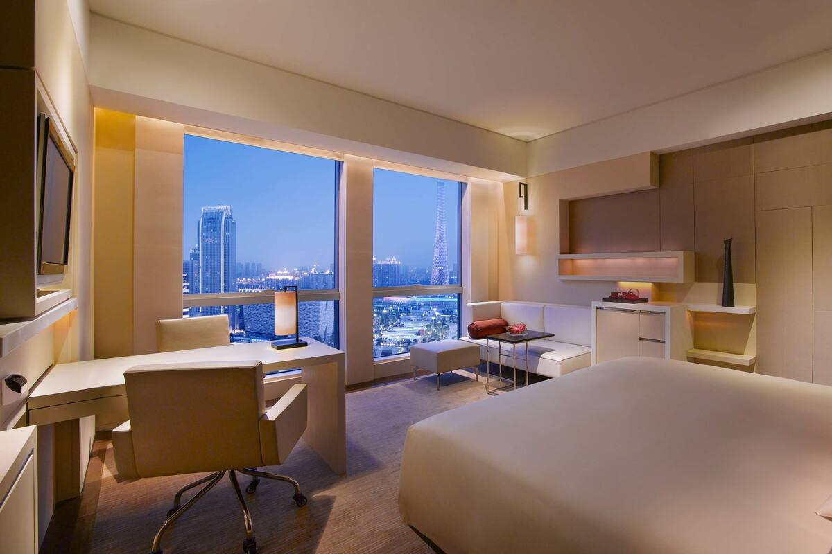 Een moderne hotelkamer met een groot bed, een bureau en een stoel. Er is uitzicht op de stad door grote ramen.