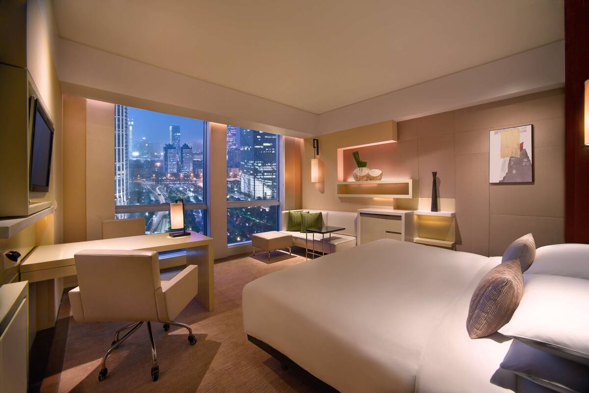 Een moderne hotelkamer met een groot bed, een bureau en een stoel. Door het raam is er uitzicht over de stad, en de kamer heeft een minimalistisch design met neutrale kleuren.