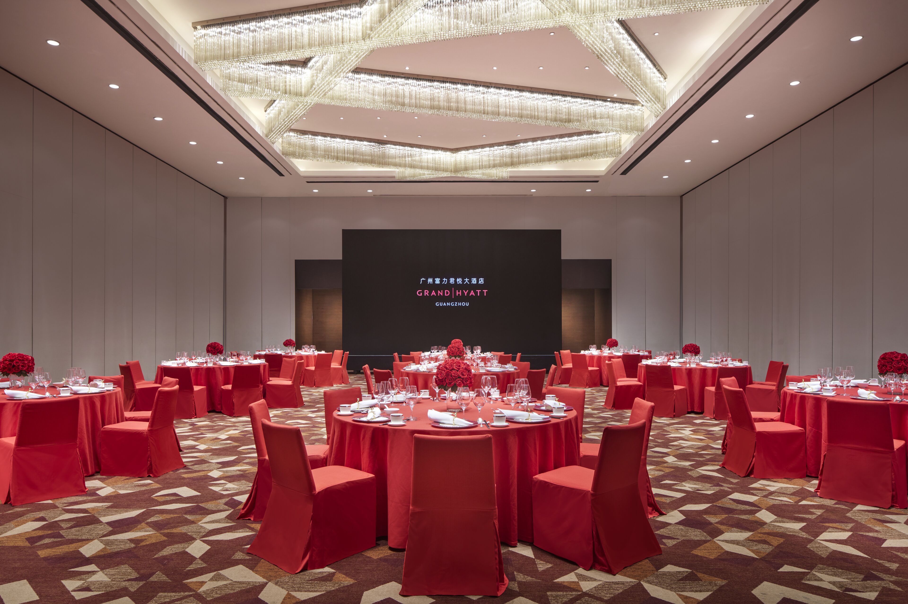 A spacious banquet hall with round tables, red tablecloths, and matching chairs. A large screen displays the hotel name.