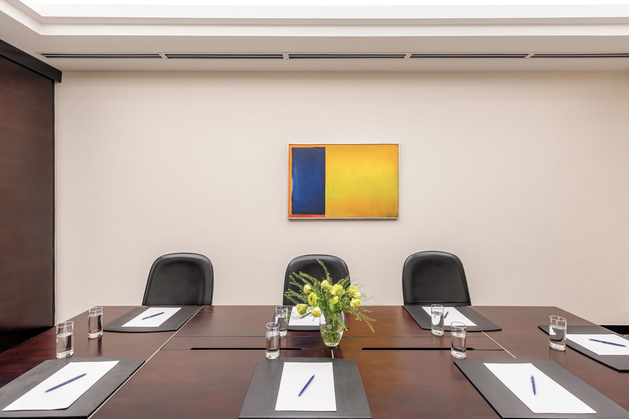 A conference room with a long table, three chairs, a vase with flowers, and a painting on the wall.