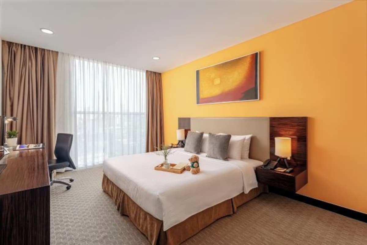 A hotel room with a large bed, a desk, a chair, a nightstand with a lamp, and a painting on the wall.