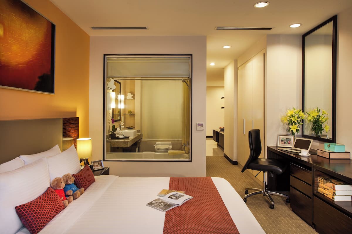 Een hotelkamer met een bed, een bureau, een stoel, een spiegel en een badkamer die zichtbaar is door een glazen wand.