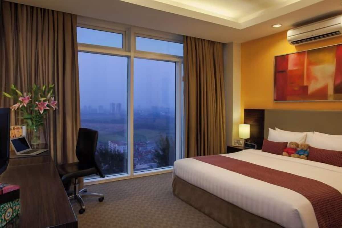 Een hotelkamer met een groot bed, een bureau met een laptop, een stoel en uitzicht op de stad door grote ramen.
