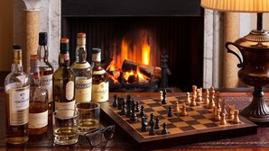 A chessboard set on a wooden table with various bottles of whisky and glasses, in front of a fireplace with a lit fire. - Burythorpe House (Malton)