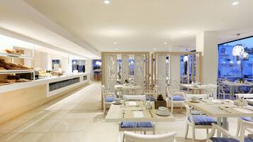 A modern restaurant with white furniture, a buffet area, and a view of the city at dusk.