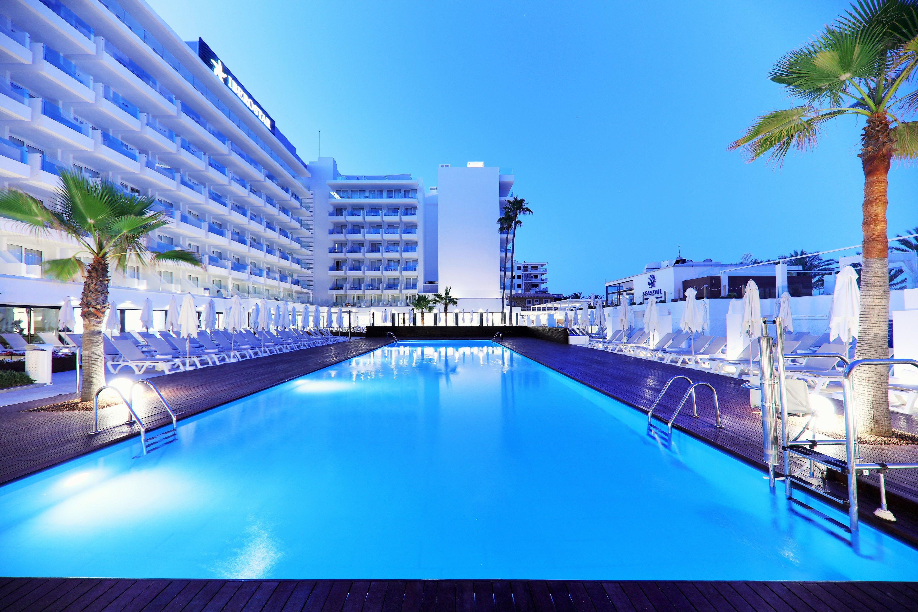Zona de piscina con tumbonas y palmeras, rodeada por un edificio de varios pisos con balcones.