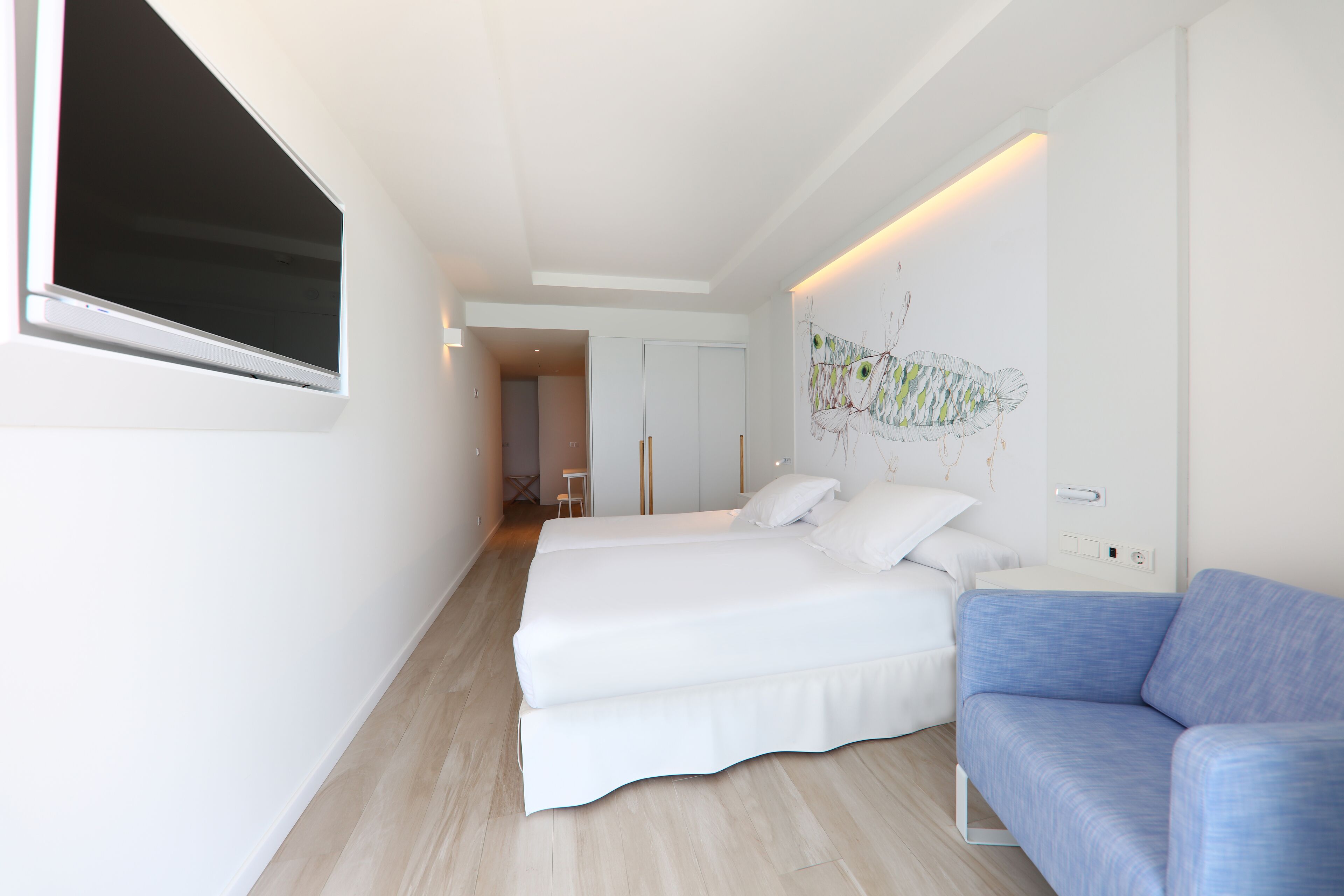 A modern hotel room with two beds, a blue sofa, a large window, and a wall-mounted artwork.