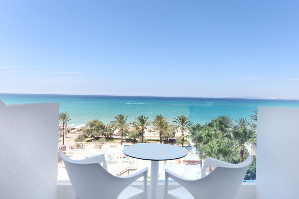 Ein Balkon mit weißem Tisch und Stühlen mit Blick auf einen Strand, Palmen und einen strahlend blauen Himmel.