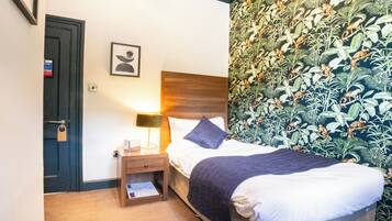 A hotel room with a bed, nightstand, and a wall with a leafy pattern.