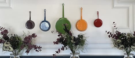A wall with several hanging pans in different colors and a fireplace with two vases of flowers.