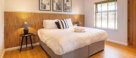 A modern bedroom with a large bed, wooden headboard, and a window with blinds.