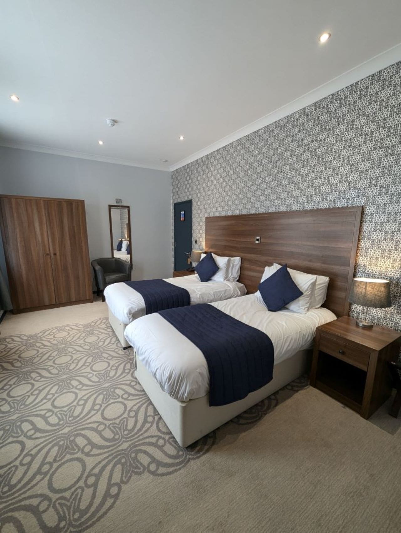 A hotel room with two beds, a wardrobe, a mirror, and a patterned carpet.