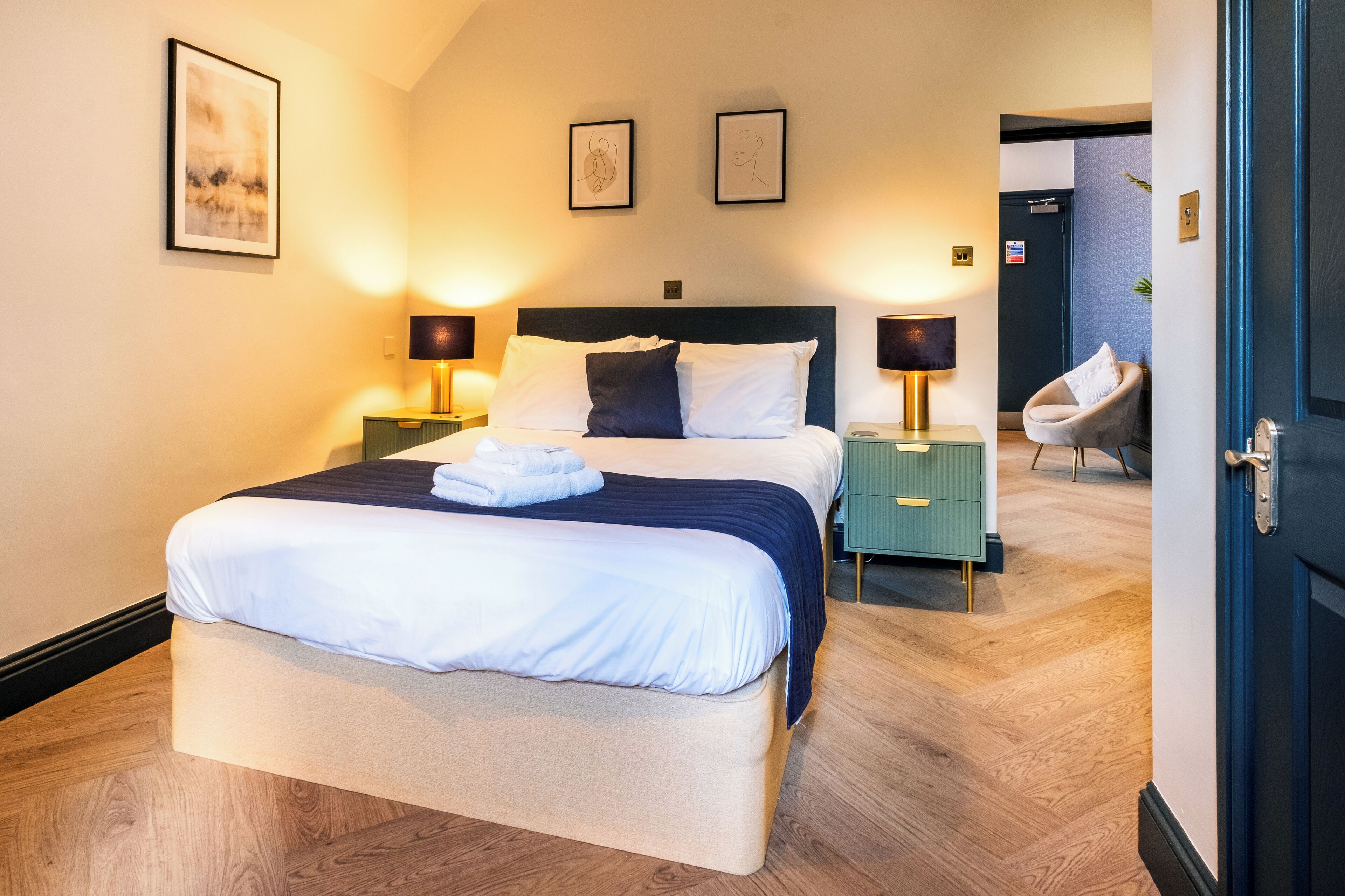A neatly made bed with white and navy bedding, two bedside lamps, a green nightstand, and a chair in a modern hotel room.