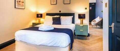 Una cama bien hecha con ropa de cama blanca y azul marino, dos lámparas de noche, una mesita de noche verde y una silla en una habitación de hotel moderna.
