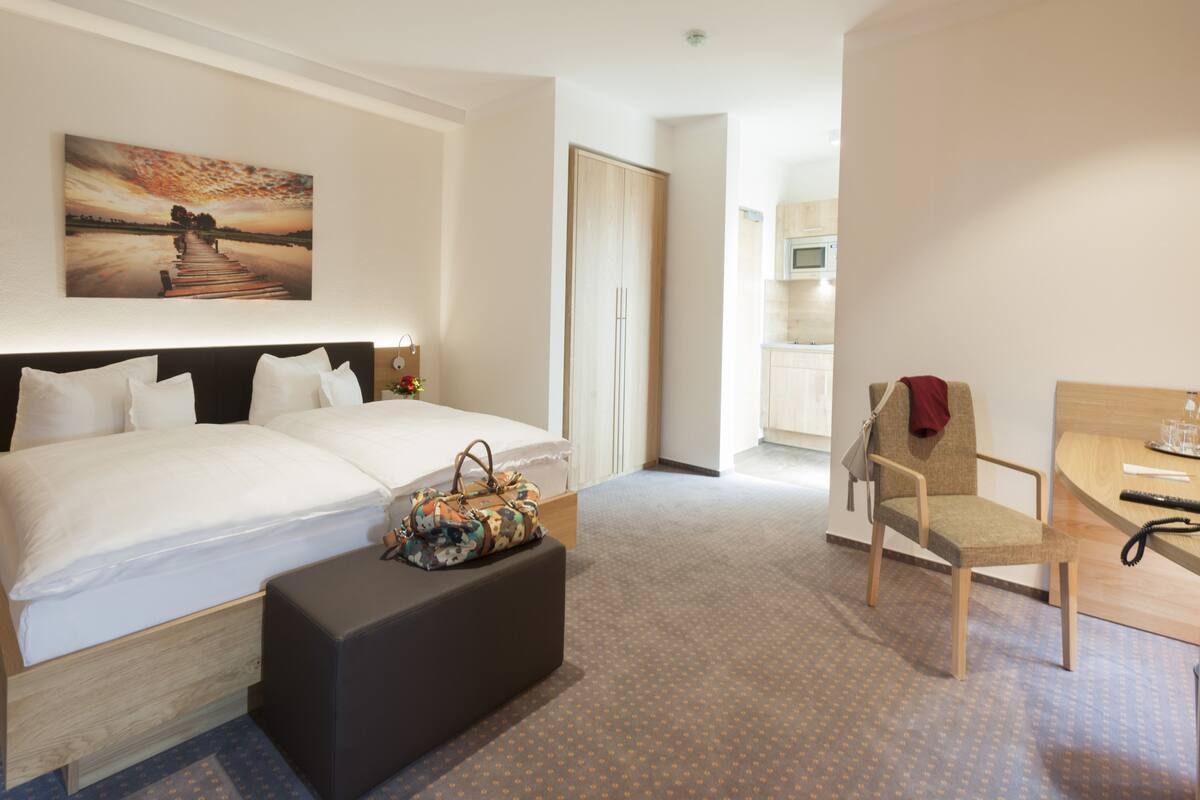 Habitación de hotel con cama, una silla, una mesita, un cuadro en la pared y una puerta que da a una cocina.