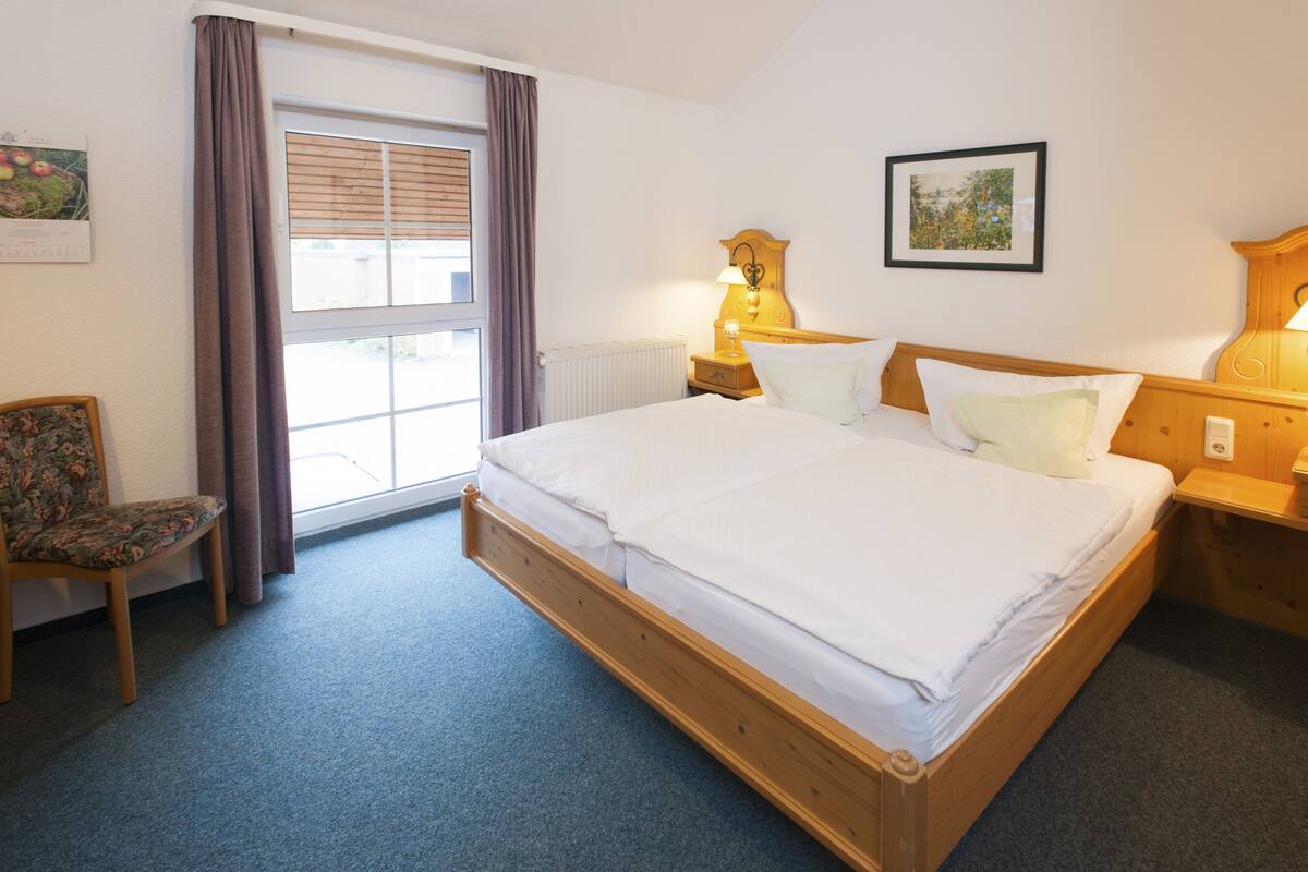 Habitación de hotel con una cama de madera, una silla, una ventana con cortinas y una lámpara fijada a la pared.