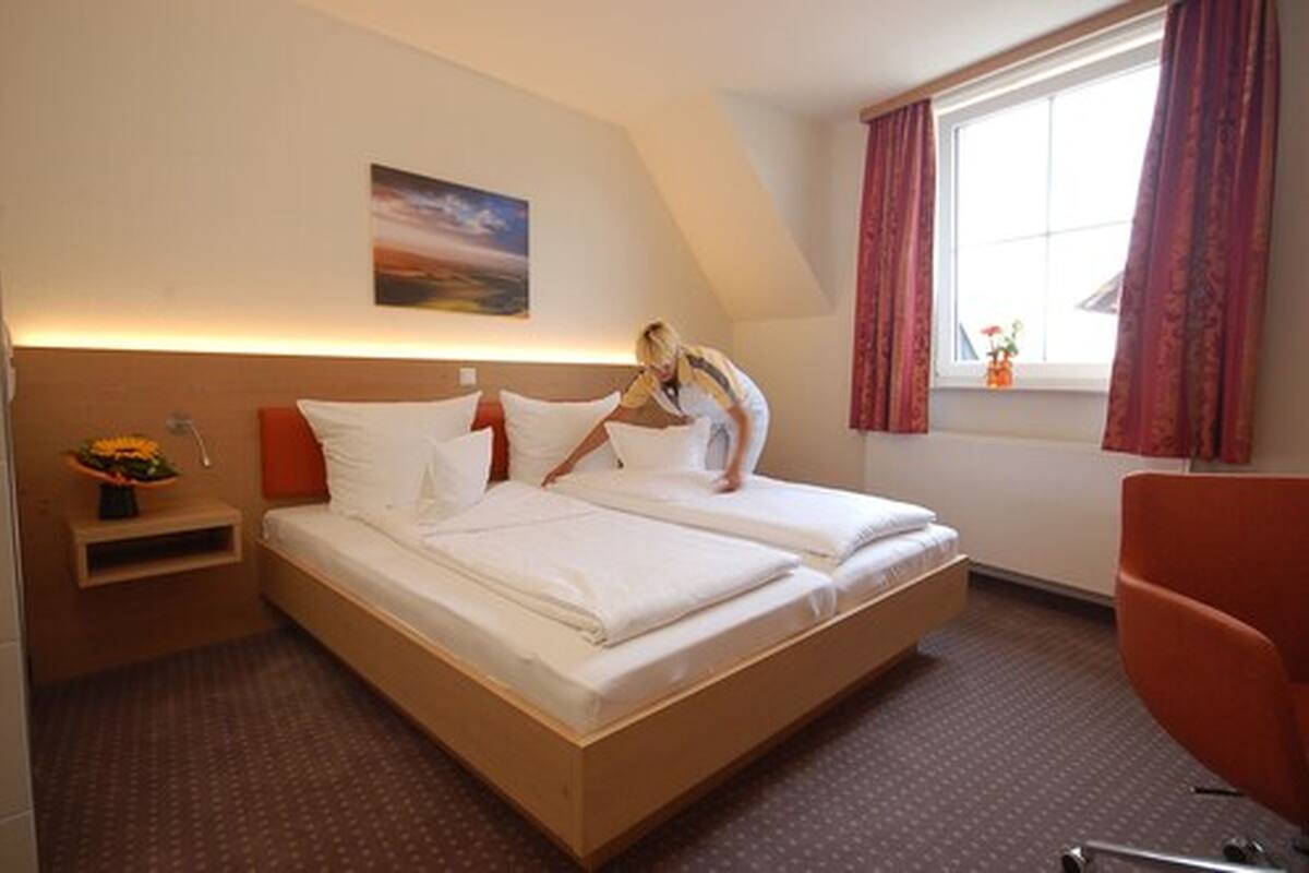 Una habitación de hotel con una cama grande, una mesita de noche con un jarrón de flores, una ventana con cortinas y un cuadro en la pared.