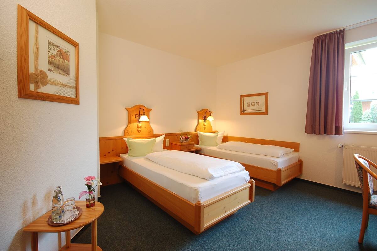 Habitación de hotel con dos camas individuales, una mesita de noche de madera con una lámpara, una pequeña mesa con un florero y botellas, y una ventana con cortinas.