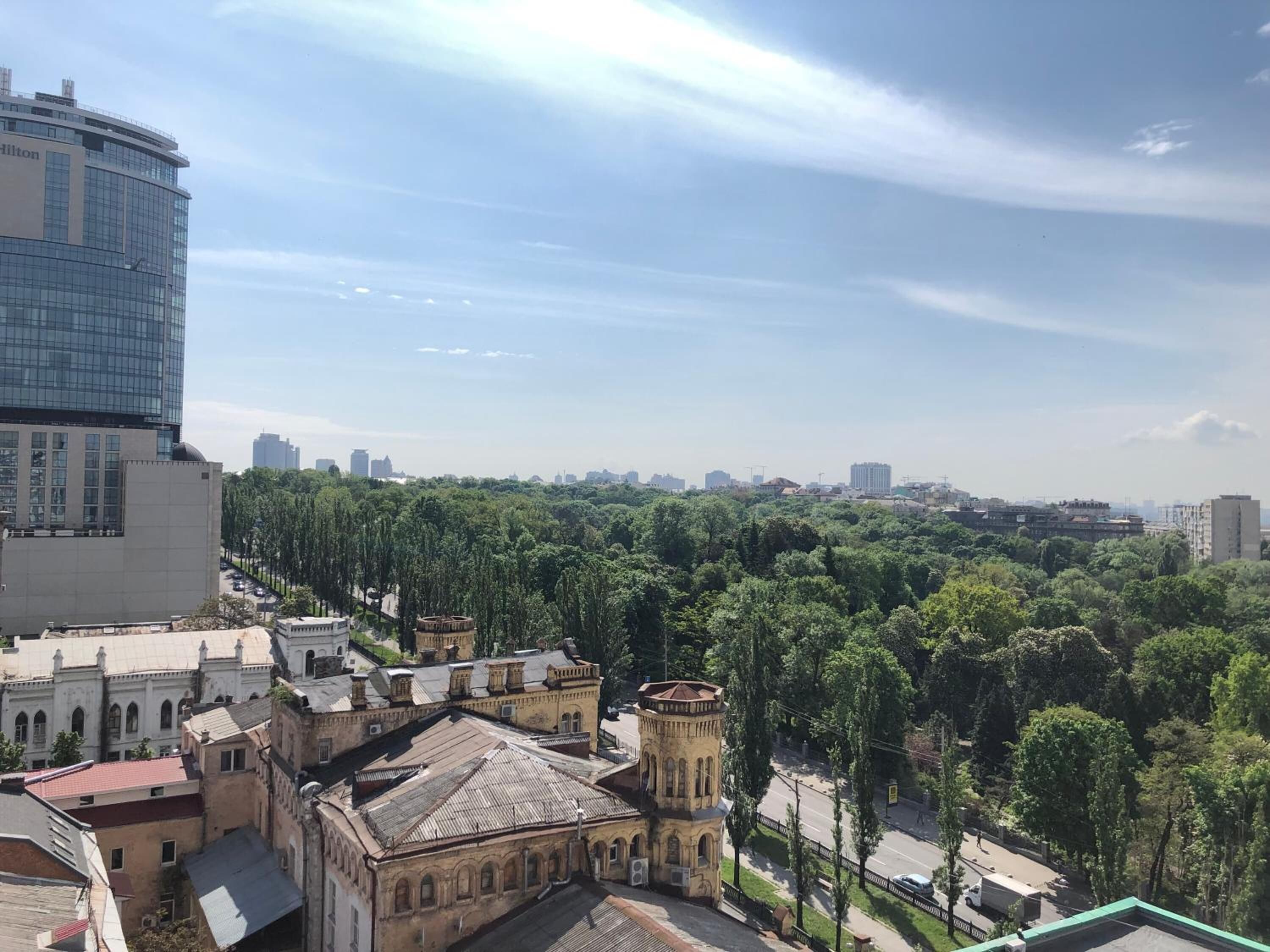 A hotel with a modern glass facade, surrounded by historic buildings and a dense tree line.