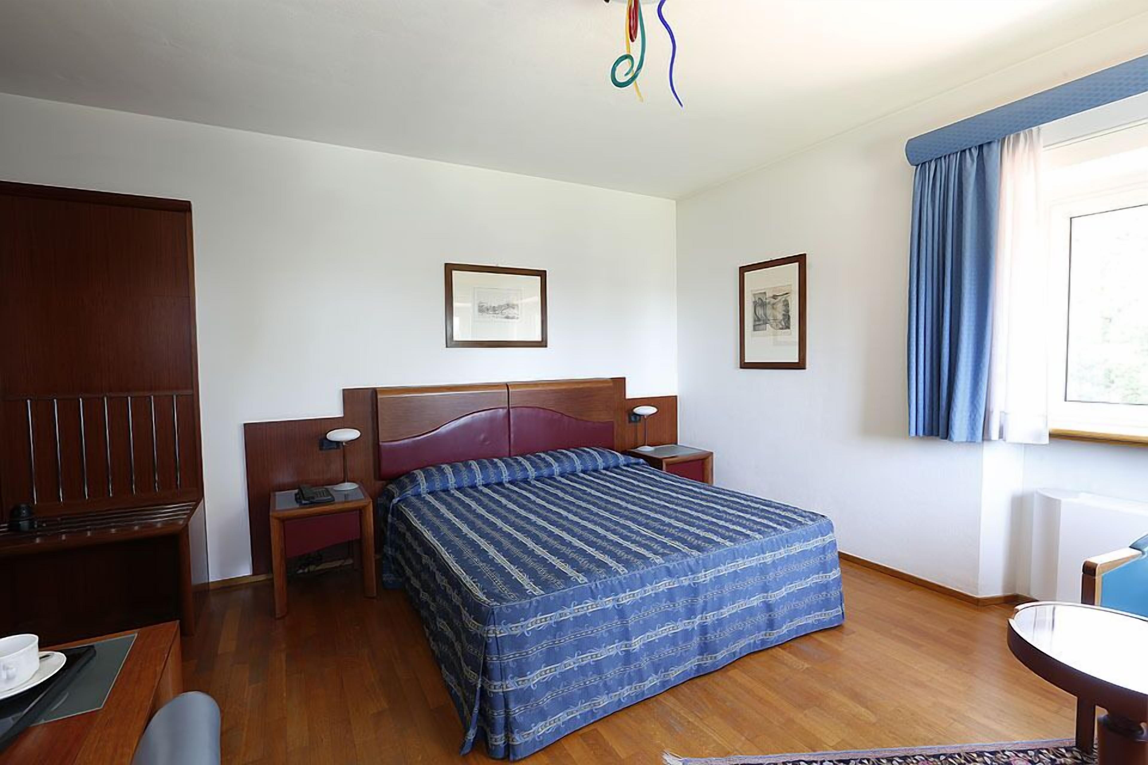 Une chambre d’hôtel avec un lit en bois, un bureau avec une chaise, une table de chevet avec une lampe et une fenêtre avec des rideaux.
