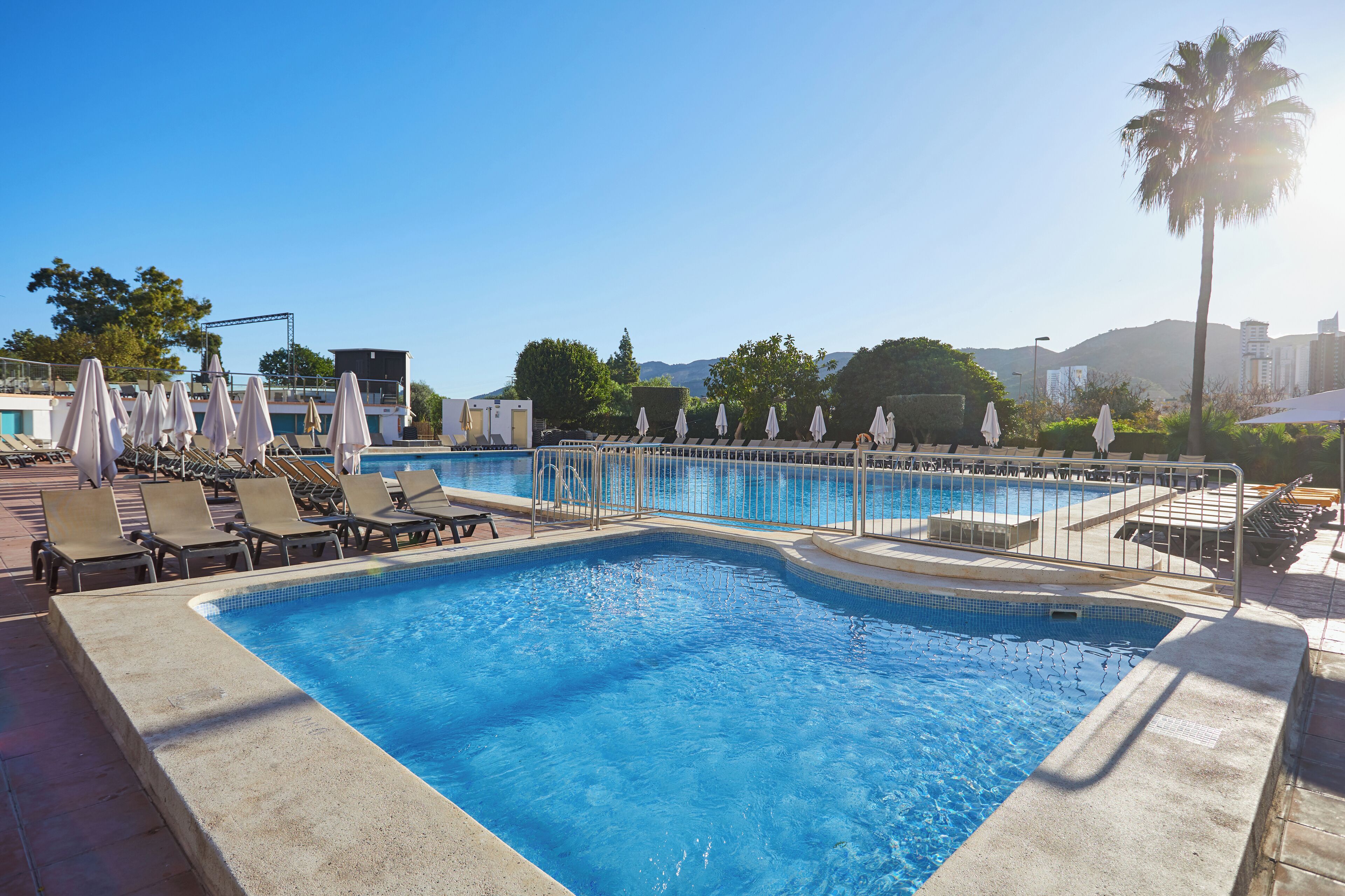 Una piscina con lettini e ombrelloni, circondata da vegetazione e edifici sullo sfondo.