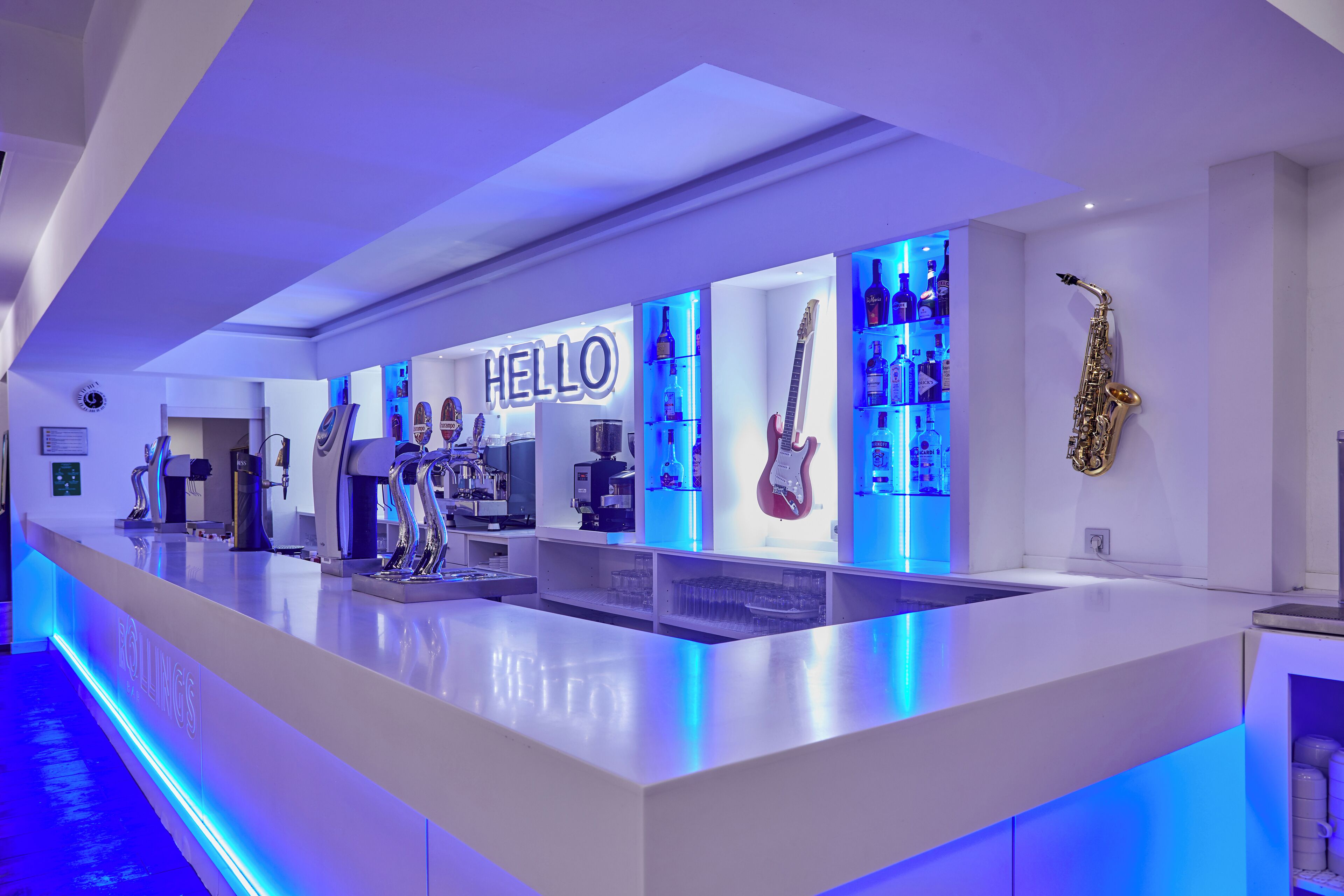 A modern bar area with a neon 'HELLO' sign, a saxophone, and various bottles.